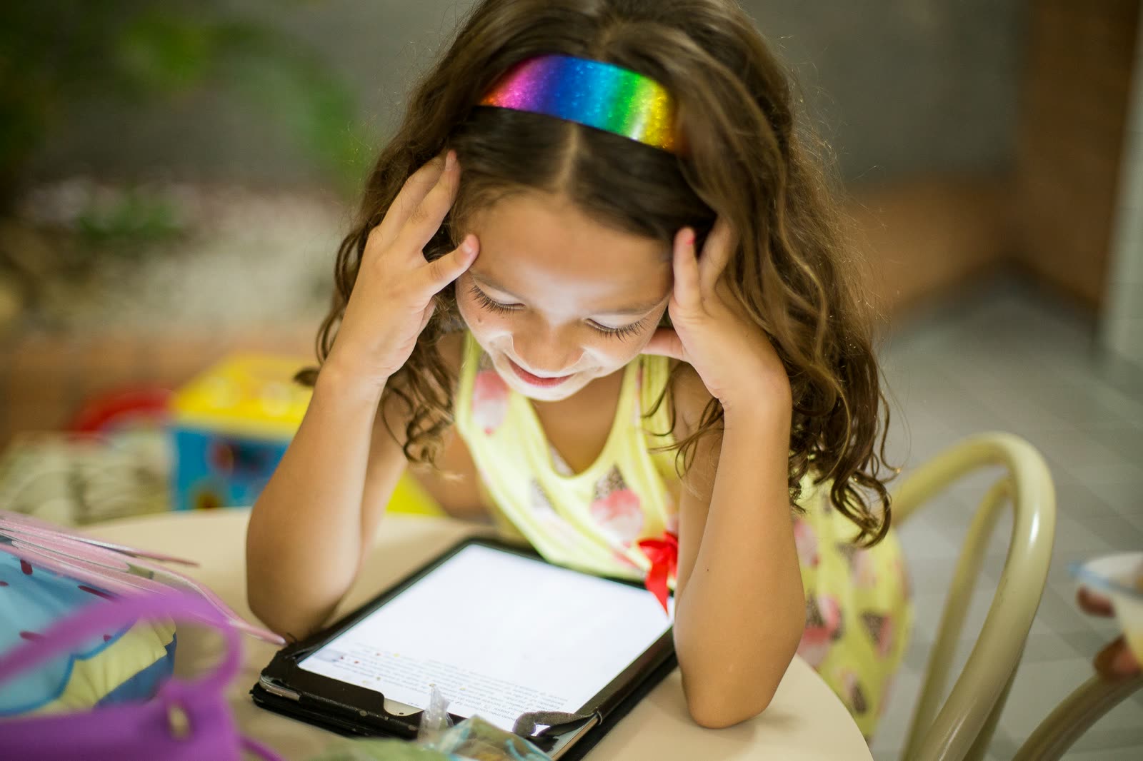 A young girl in a rainbow headband reading on a tablet at a table